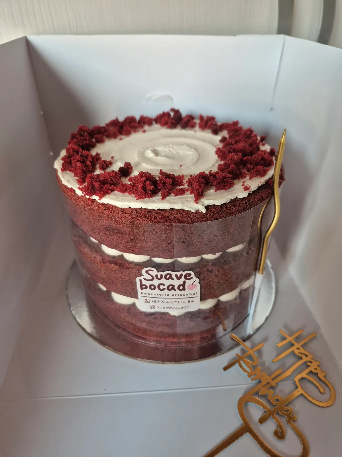 Torta de bizcocho rojo con capas de crema blanca y migas rojas encima, dentro de su caja