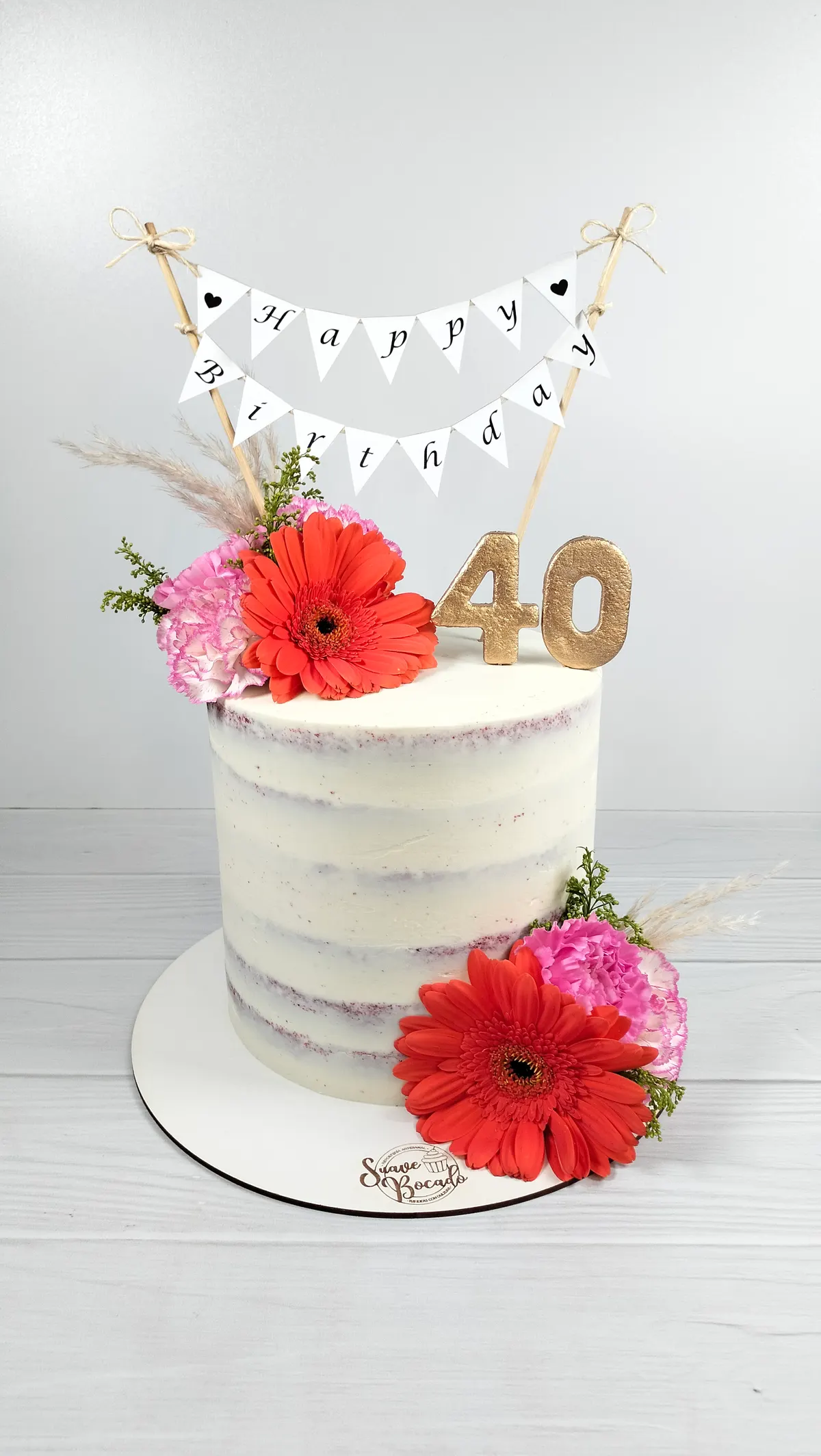 Torta blanca semidesnuda con gerberas rojas y rosadas, un banderín de Happy Birthday y un 40 dorado