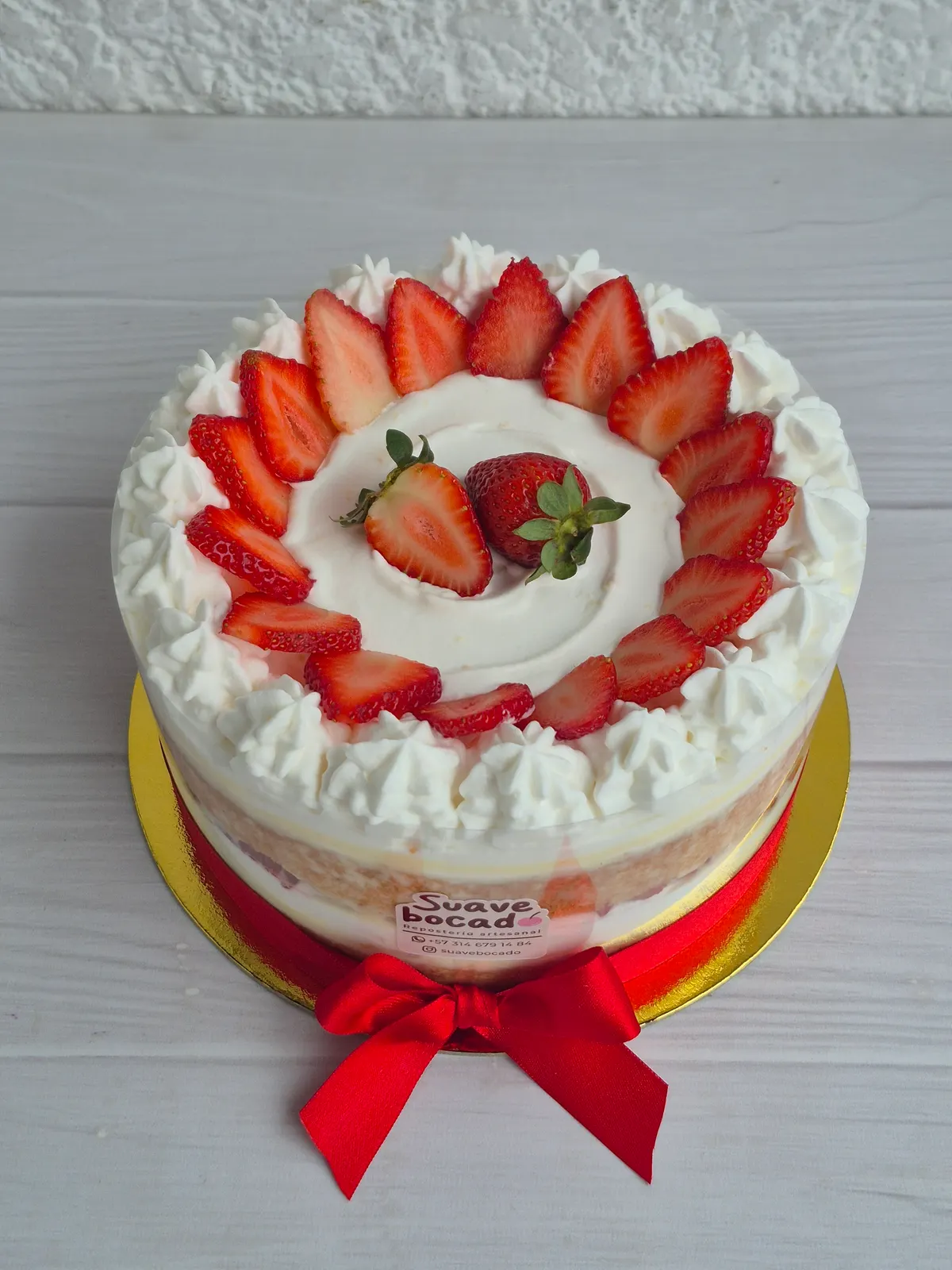 Torta de crema blanca con un círculo de fresas encima y un lazo rojo alrededor