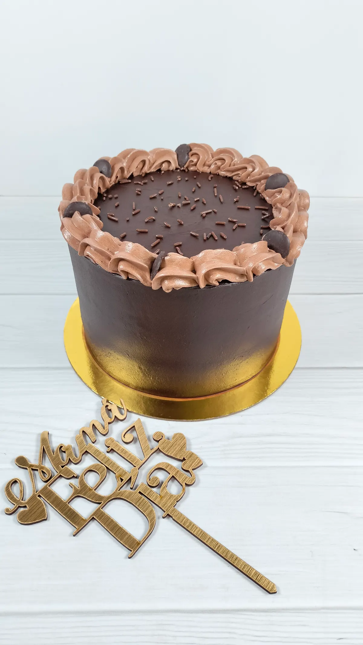 Torta cubierta de chocolate con borde de crema de chocolate, junto a un topper de Mamá feliz día