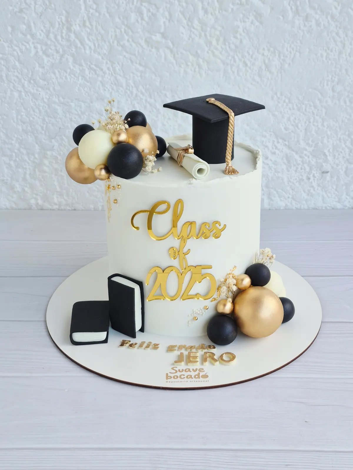 Torta de grado blanca con birrete, diploma, esferas negras y doradas y un topper de Class of 2025
