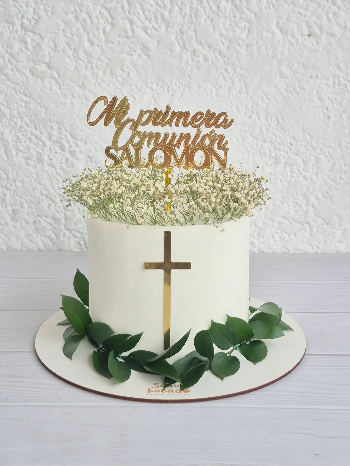 Torta de primera comunión blanca con una cruz dorada, hojas verdes y flores de nube encima