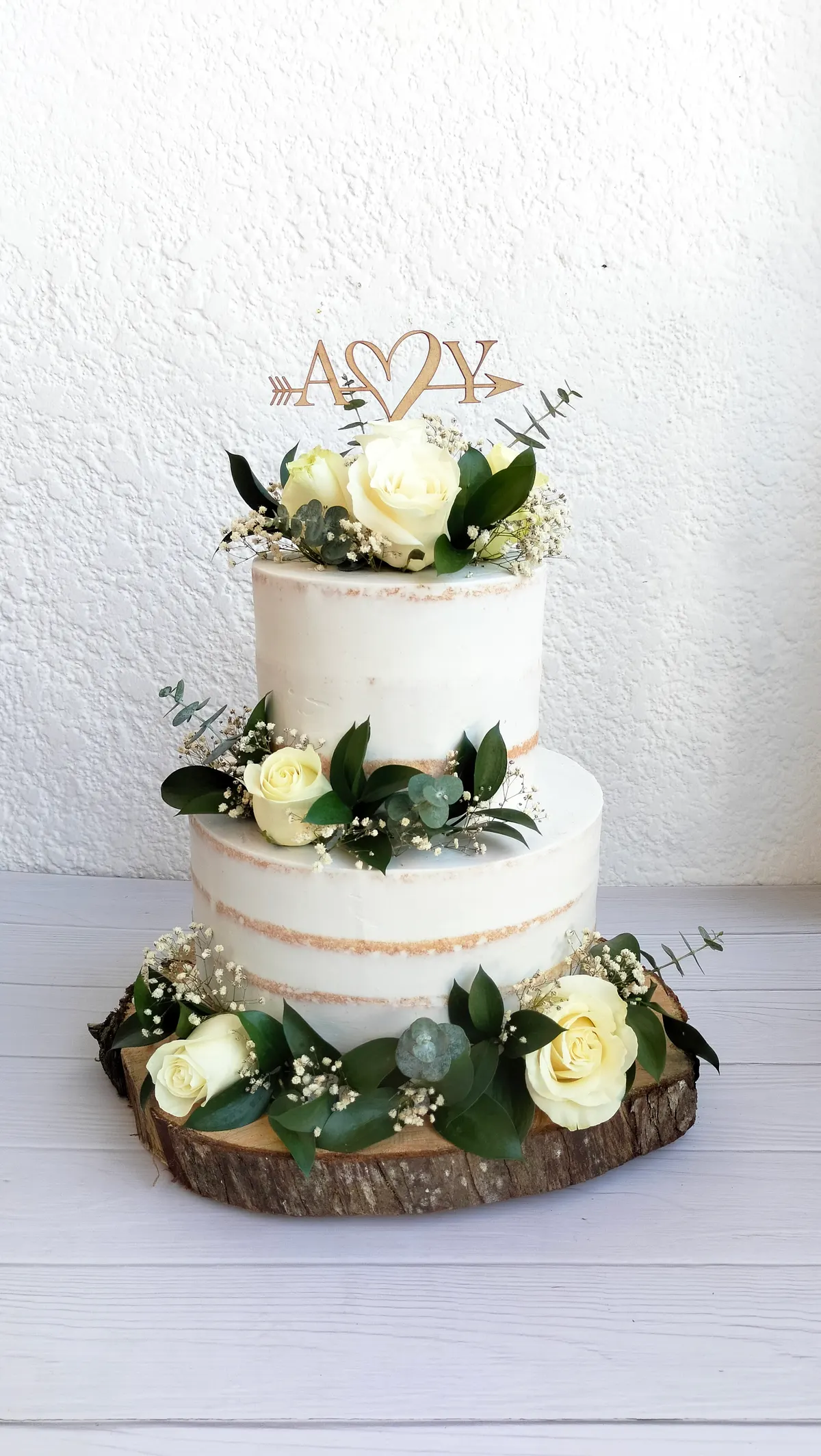 Torta de matrimonio de dos pisos con rosas blancas, hojas verdes y las iniciales A y Y, sobre un tronco