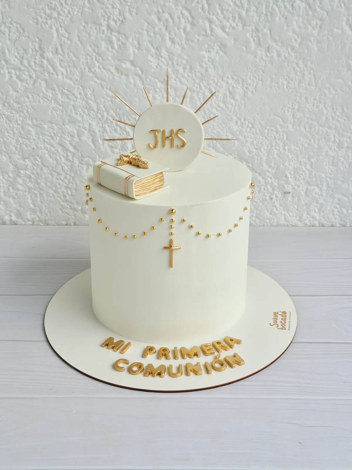 Torta de primera comunión blanca con una hostia JHS entre rayos dorados, un libro, un rosario de perlas y el mensaje Mi primera comunión