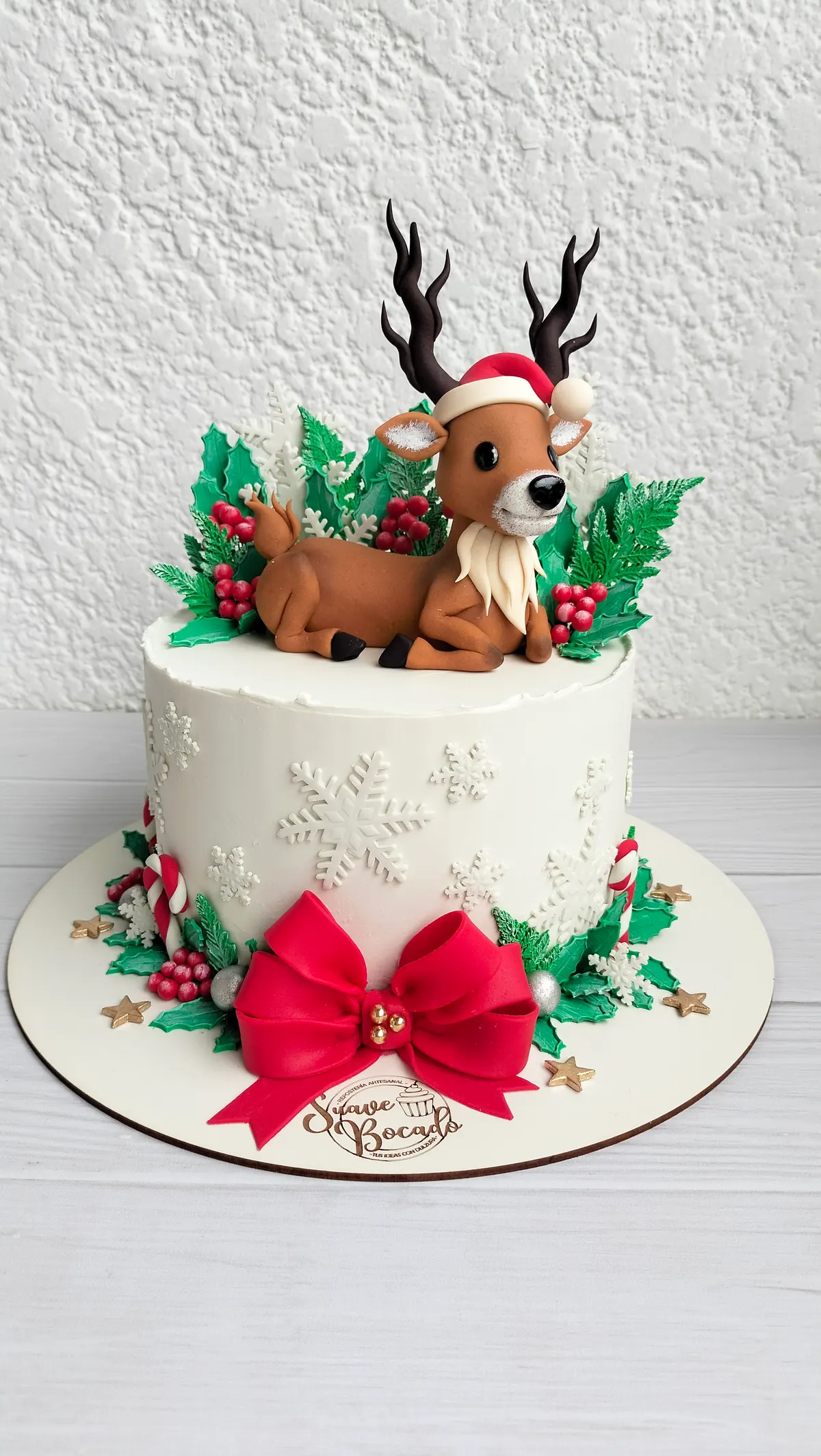 Torta de Navidad blanca con copos de nieve, un reno con gorro de Papá Noel, hojas de acebo y un lazo rojo